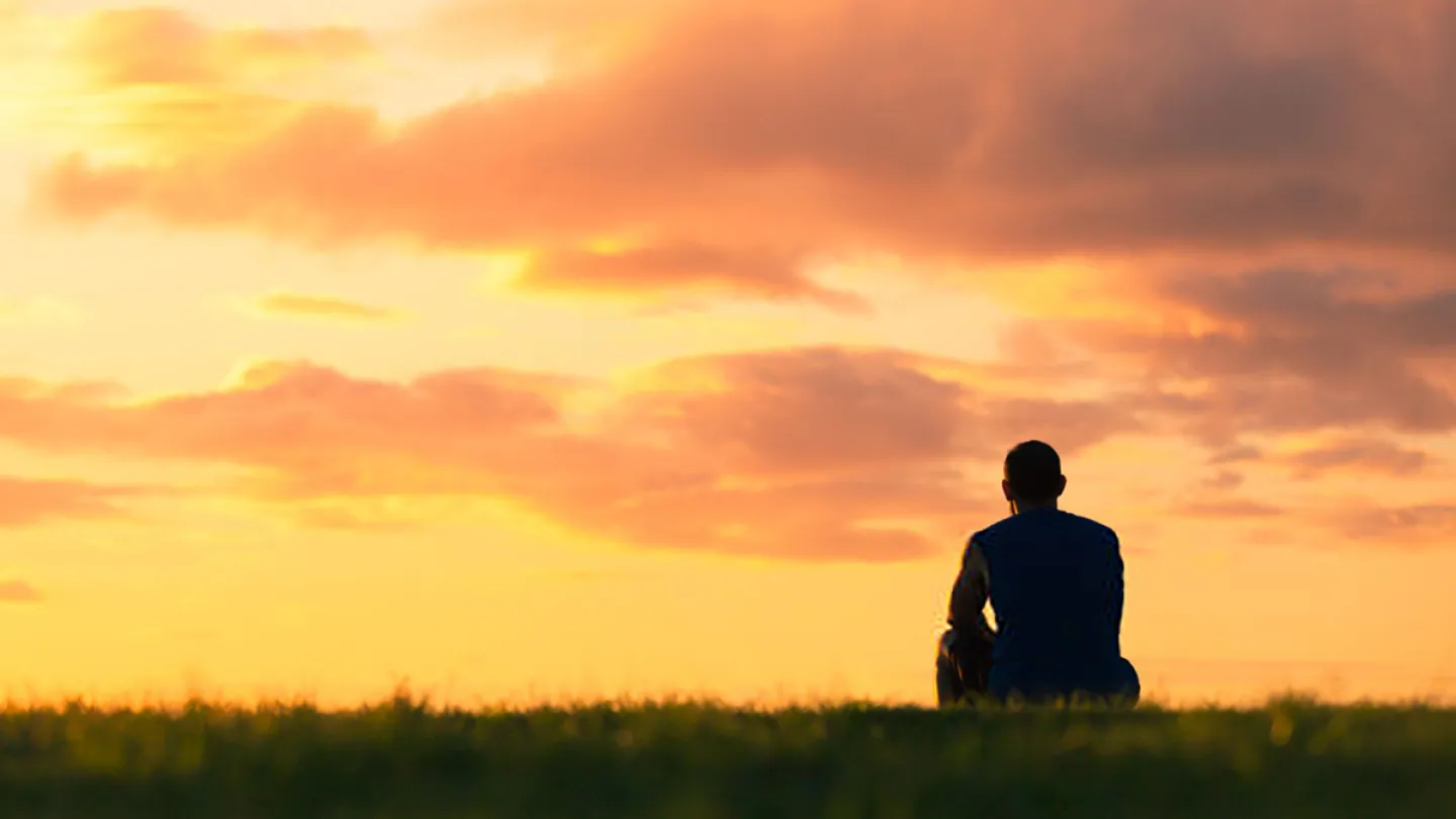 Person sitting alone on grass, looking out at a sunset sky