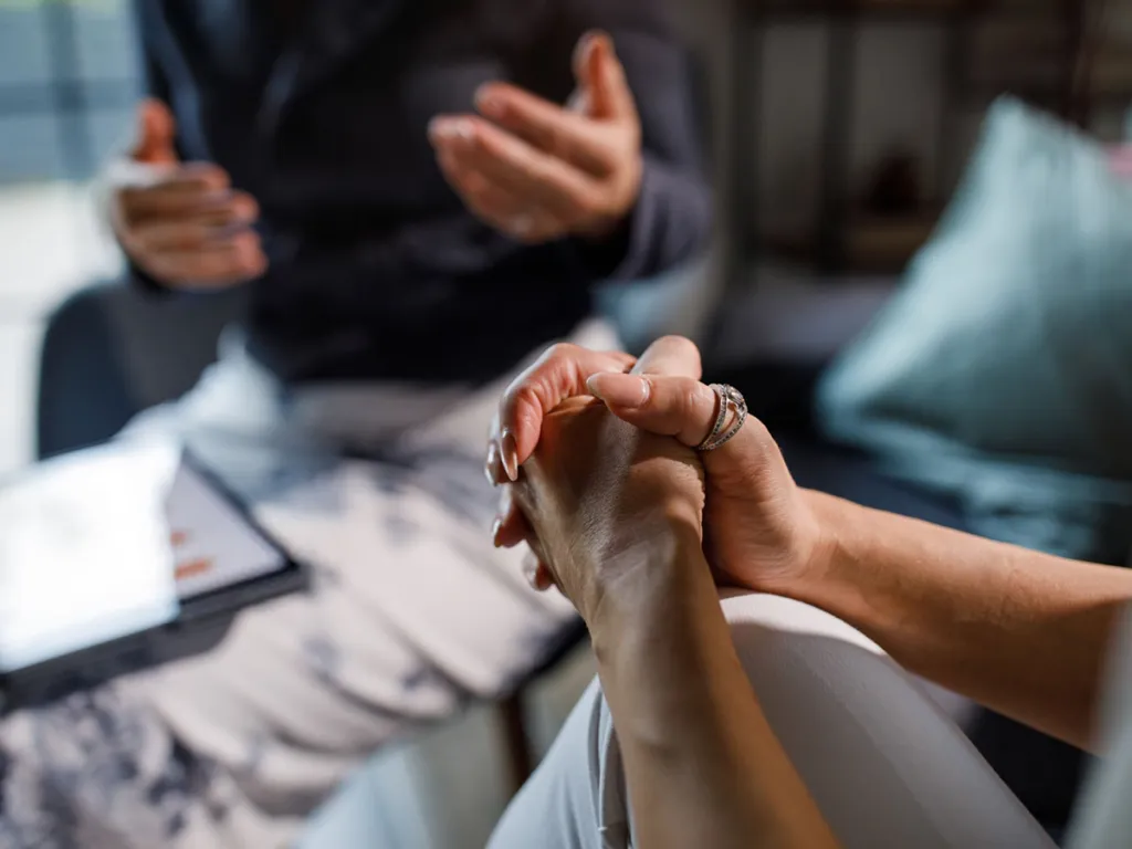 Close-up of one person holding another’s hands in a supportive conversation, with a third person gesturing in the background