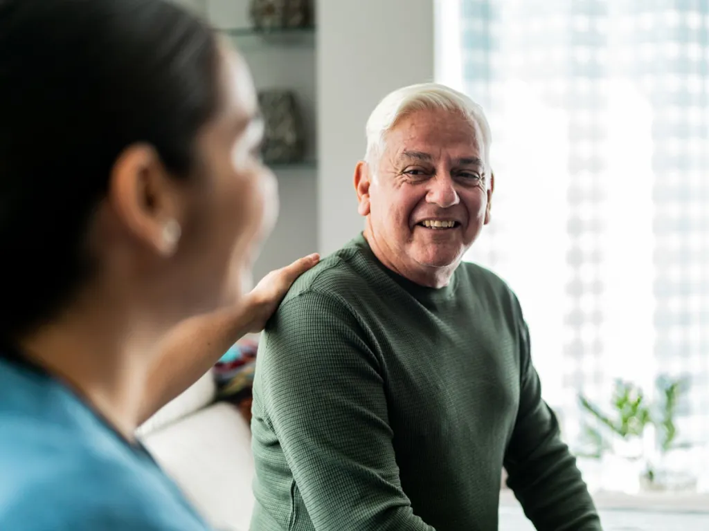 Older man smiling while a woman places a reassuring hand on his shoulder during a conversation at home