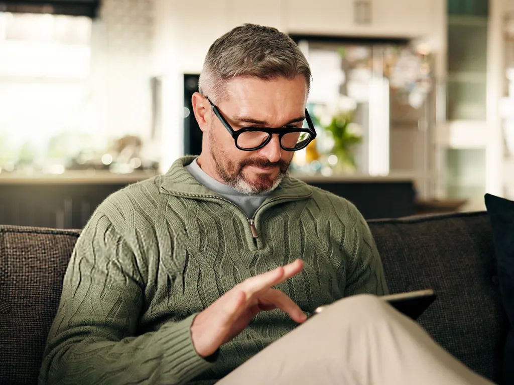 Man wearing glasses sitting on a sofa, using a tablet in a relaxed home setting