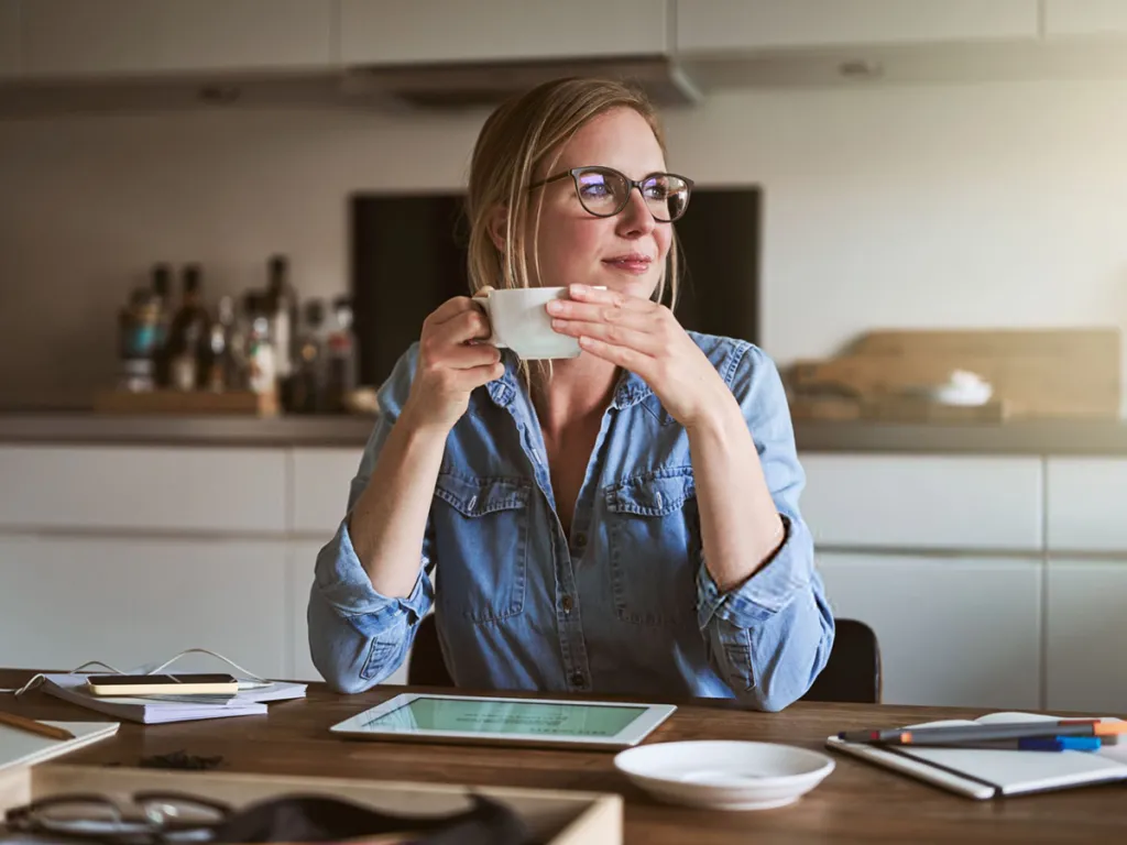 Woman sitting at a kitchen table holding a cup of coffee, with a tablet and notebook in front of her