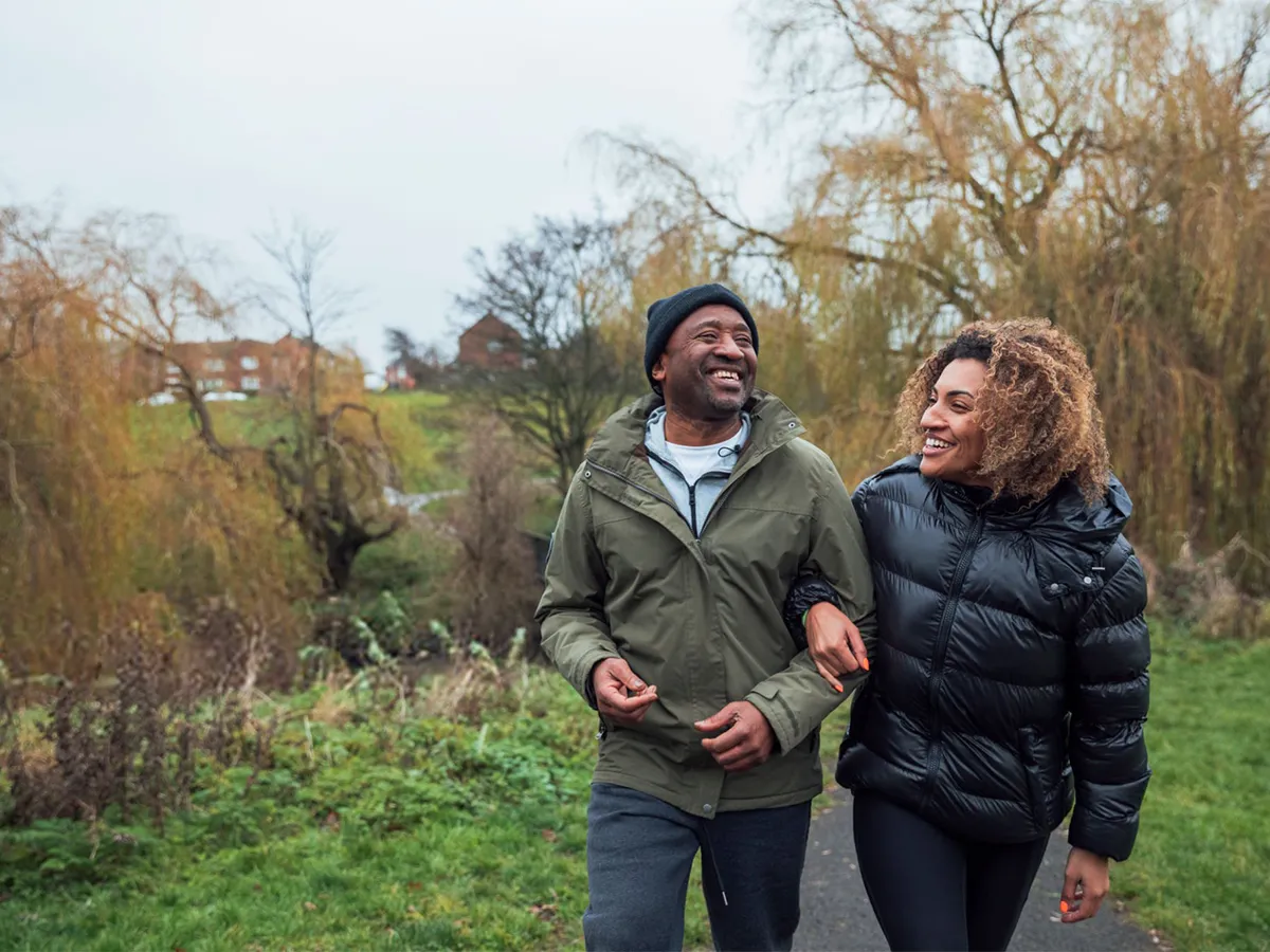 Two people walking together outdoors on a path, smiling and talking, with trees and houses in the background