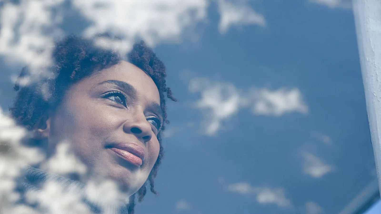 Woman looking out through a window, reflected with clouds in the glass